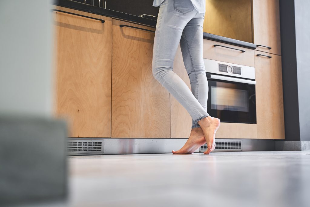 What's the Best Flooring for a Kitchen Remodel? 3 Woman with bare feet in jeans standing in a kitchen next to wood grain cabinets and concrete color flooring.